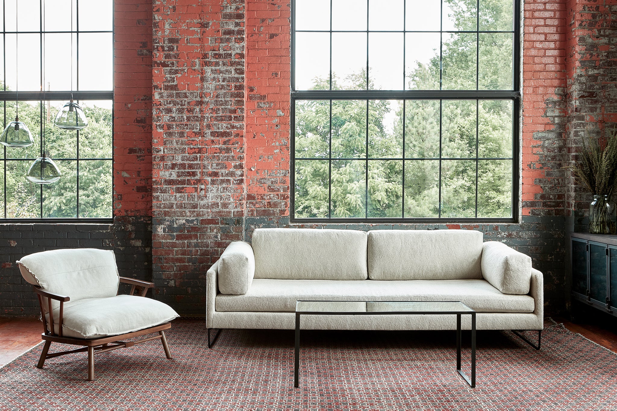  A sofa with 2 back cushions and side pillows in a textured neutral fabric in front of an orange vintage brick wall with 3 smoke glass lights and large industrial windows. There is a chair with a wood frame and a neutral linen cushion on the left side. In front of the sofa, there is a metal and glass coffee table. On the right there is a metal and glass credenza with a large flower arrangement. The rug is dark red and grey with a geometric pattern. Photographed in Segura Natural. 
