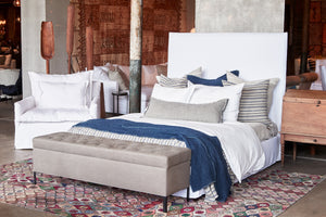  The April Tall Bed is in a white slipcover, there are many decorative pillows on it, a white duvet cover and a navy blue throw. The Jaxon Chest at the end of the bed is in grey linen. To the left of the bed, there is a white slipcovered armchair. The rug is multicolored with a flower pattern. Photographed in Logan White. 