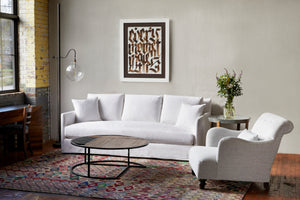  Living room setting with large windows. White slipcovered sofa with white Acacia Chair and round wooden coffee table. Floor lamp on left side of sofa and art work on wall.  