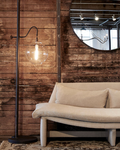 Metal floor lamp with clear circular pendant next to a sofa with neutral fabric sitting in front of a wood wall. 