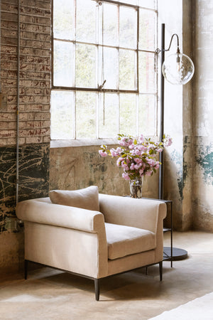  Upholstered chair in neutral fabric placed against brick wall with large window. Metal floor lamp placed on right side of upholstered chair. 