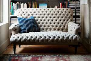  The sofa is a small scale piece with a lot of tufting, in a neutral color linen, with a blus striped pillow on the right side. The sofa is in front of a book shelf and has a vintage blue and red rug in front. Photographed in Vintage Flax. 