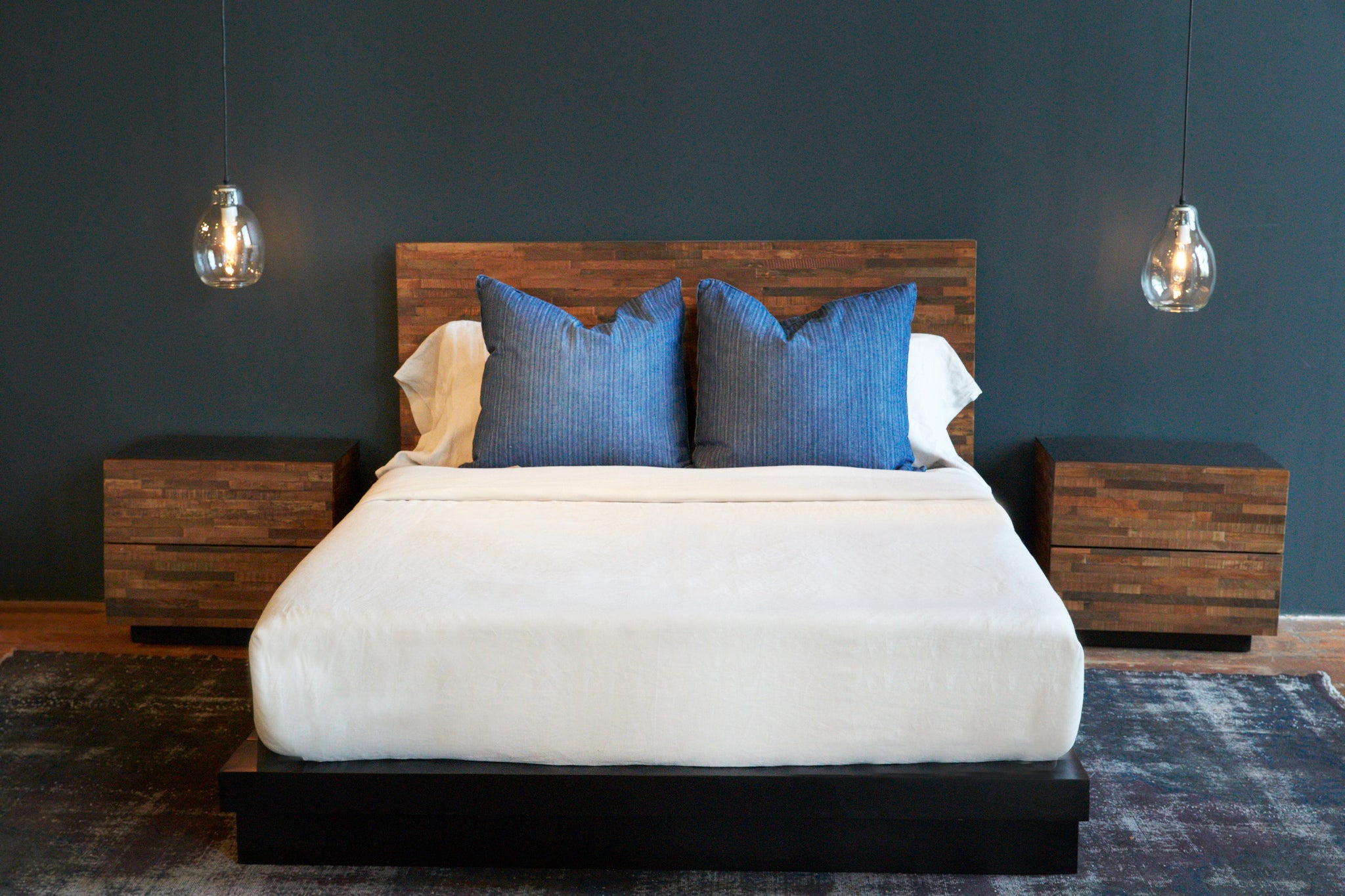  Bedroom setting. Wood platform bed  with white bedding and blue pillows with wood night stands against a blue wall and clear oval pendants hanging above each nightstand.  