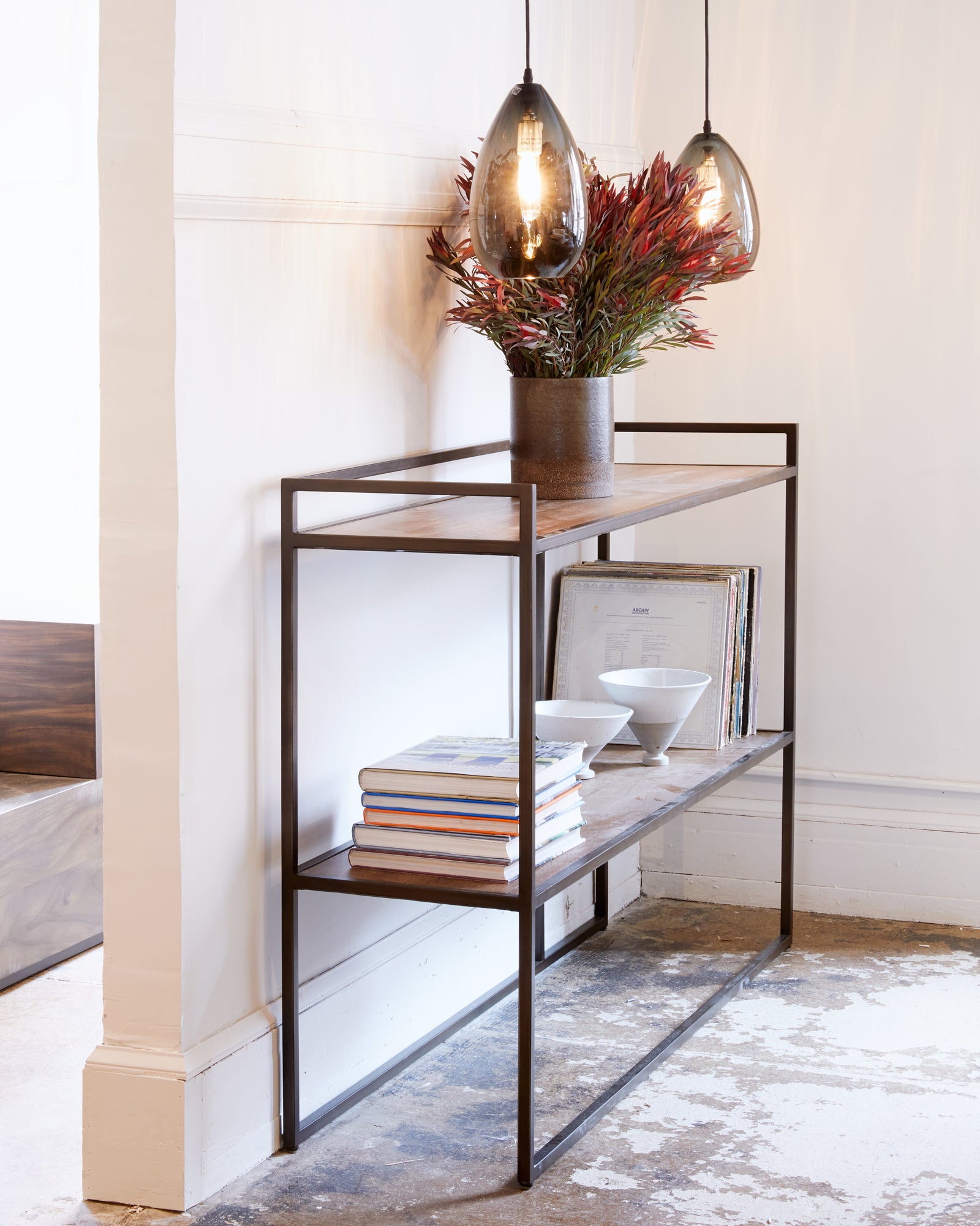  Wood console with books, potter and flower vase placed against white wall. Oval hanging pendants in smoke color hanging above console.  