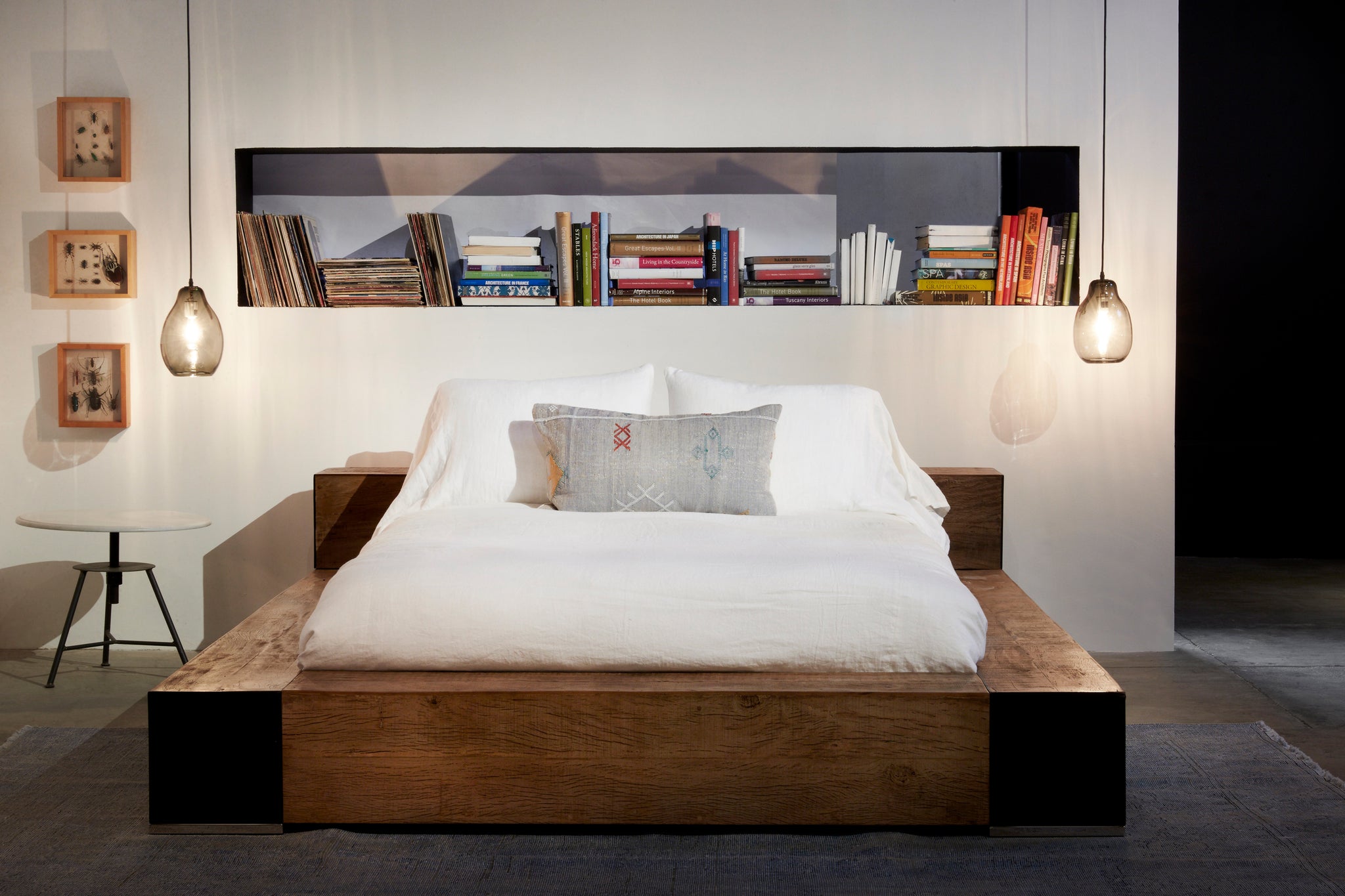  Bedroom setting. Wood platform bed with white bedding against white wall with book nook and smoke colored oval pendants hanging on either side of bed.  