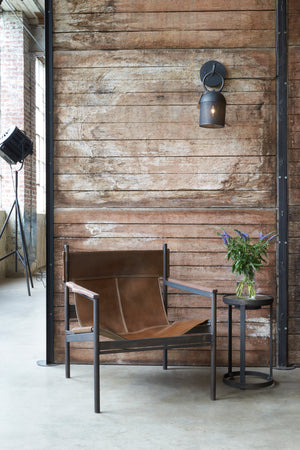  Chair in Vachetta Coffee next to side table. In the background is a wood wall with a lighting piece above. Photographed in Vachetta Coffee. 