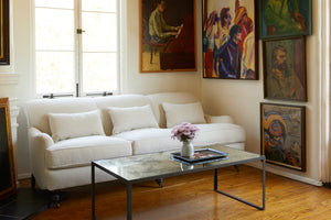  The sofa is in a neutral linen color, with 3 small lumbar pillows. It is in a small room with wood floors, in front of a window. On the right there are 5 vintage paintings, in front there is a rectangular coffee table with a vintage mirror top. There is a blue vase with purple flowers on a black tray. Photographed in Brevard Birch. 