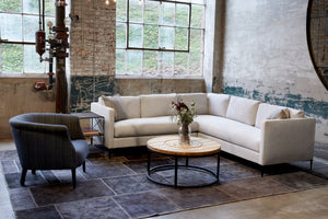  The Benedict 2 arm sectional is in a light grey fabric in the corner of an industrial looking showroom with aged cement walls and a large window. A round coffee table with a vintage wood top is in front and flowers on top. A dark grey striped Emily chair is on the left of the sectional. Grey patchwork rug on the floor. Photographed in Navedo Gravel. 