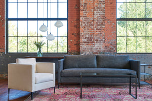  Lester Charcoal Benedict Sofa in dark fabric with the Benedict Chair in a loft with large windows and 3 white glass pendant lights. Photographed in Lester Charcoal. 