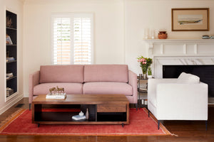  The sofa is in a pink fabric, the chair is on the right side in a white fabric. The room is bright with white walls, there is a bookshelf on the left side, the inside is painted black. On the right of the photo, there is a fireplace with some objects on top and a vintage painting on the wall.A rectangular wood coffee table in in front in the sofa and chair. There is a red rug, a metal side table on the right of the sofa with a vase on top with pink and green flowers. Photographed in Molino Blush. 