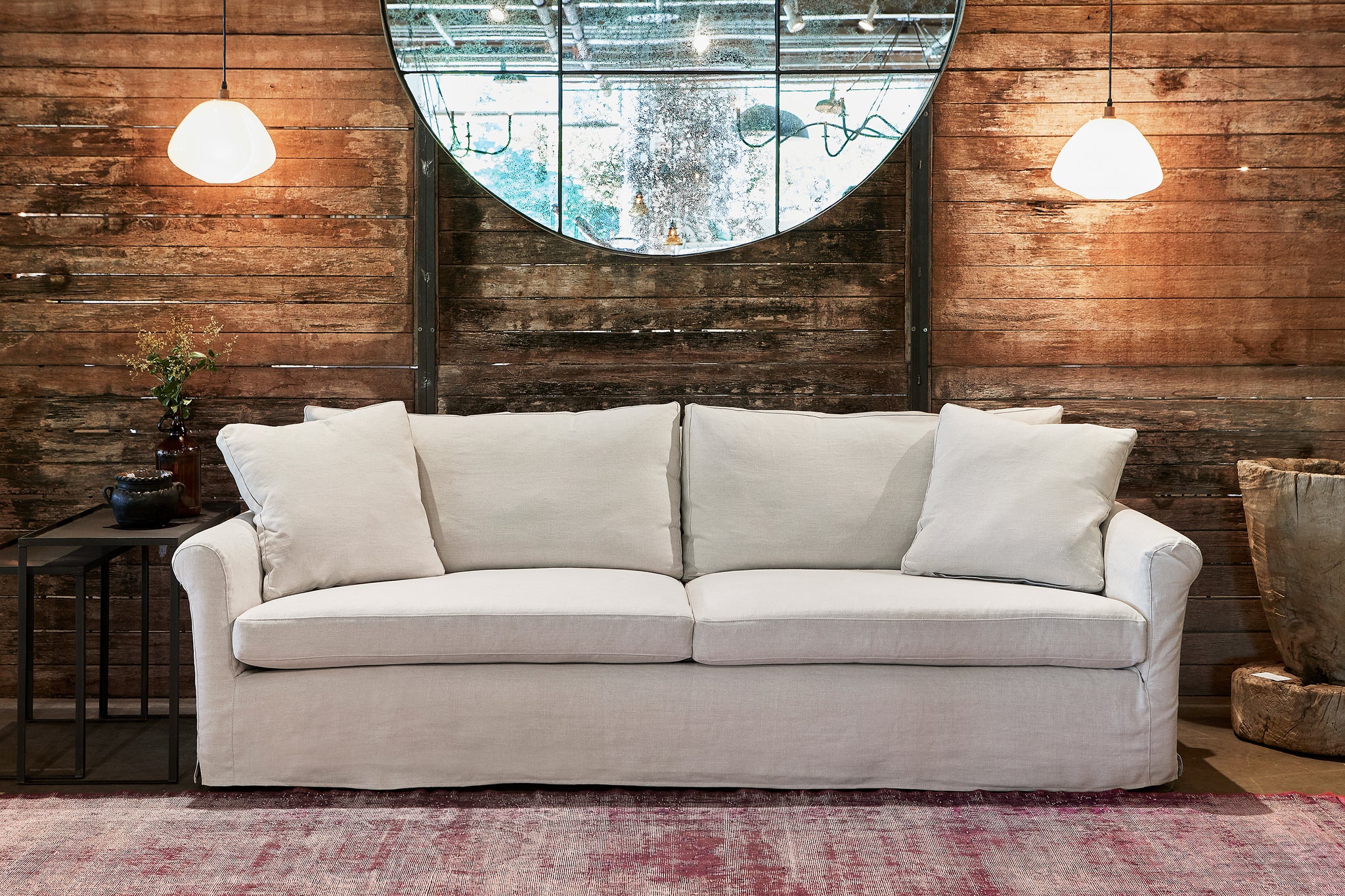  Light colored slipcovered sofa in front of a dark wood wall with 2 white lighting pendants and a large round mirror. Photographed in Noah Bone. 