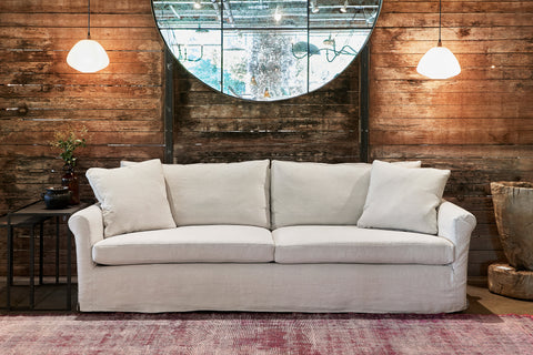 Light colored slipcovered sofa in front of a dark wood wall with 2 white lighting pendants and a large round mirror. Photographed in Noah Bone.