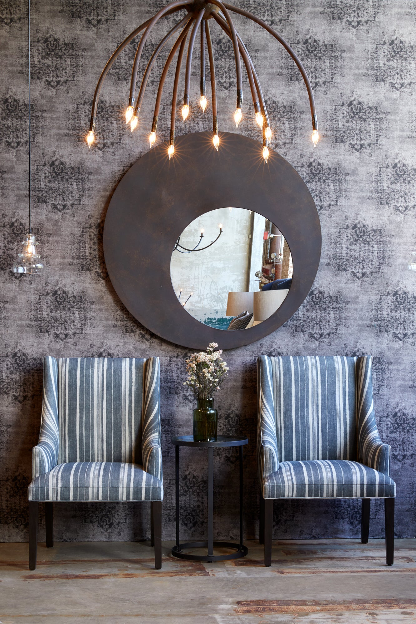 Blossom chandelier in black finish hanging above two upholstered chairs in striped blue fabric with round metal side table placed in front of upholstered wall in a grey/purple printed fabric and a round metal wall mirror.  