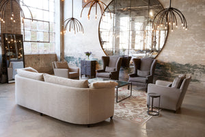  Large room with distressed brick walls and large windows. Three blossom chandeliers in black finish hanging above living room setting with a tan upholstered sofa, two grey upholstered wing back chairs, two upholstered chairs in grey fabric, metal coffee table with mirror top, floor mirror with black metal frame and a large round wall mirror.  