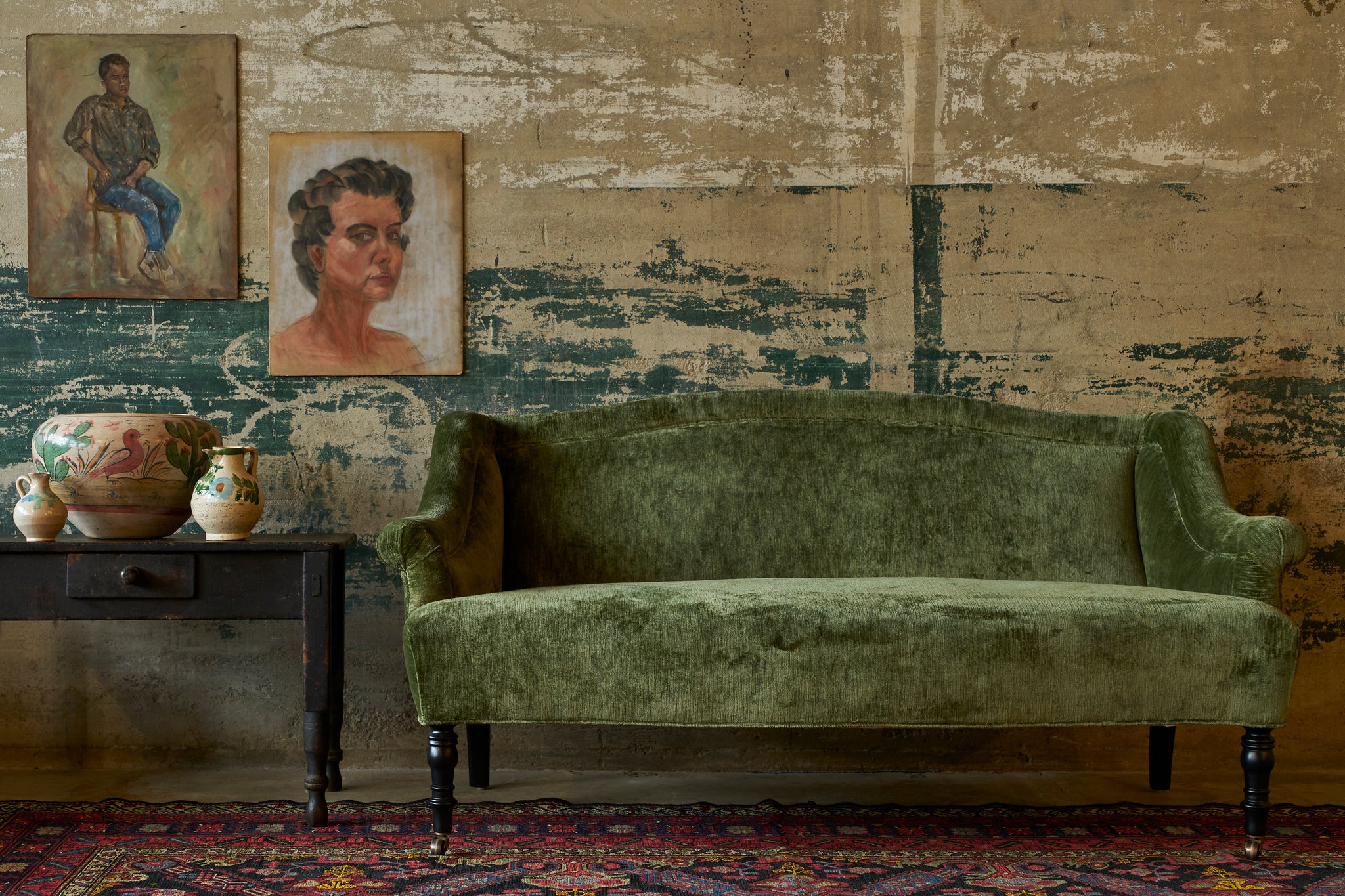  Green velvet loveseat in front of a wall with old painting next to a wood table with painted pottery. Photographed in Velluto Olive. 