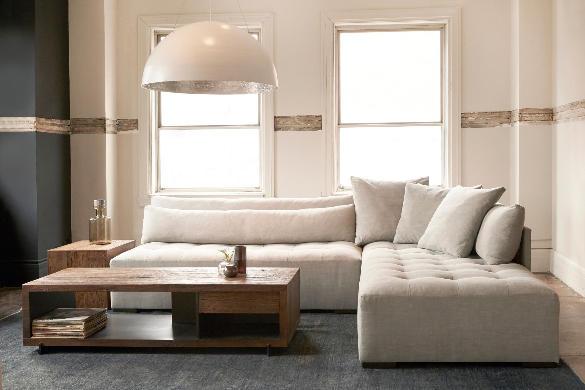  Tufted sectional upholstered in a neutral fabric with a wood coffee table placed in front of it and a white circular pendant hanging above it. Setting is placed in a white room with two large windows and sectional is placed on top of a grey rug.  
