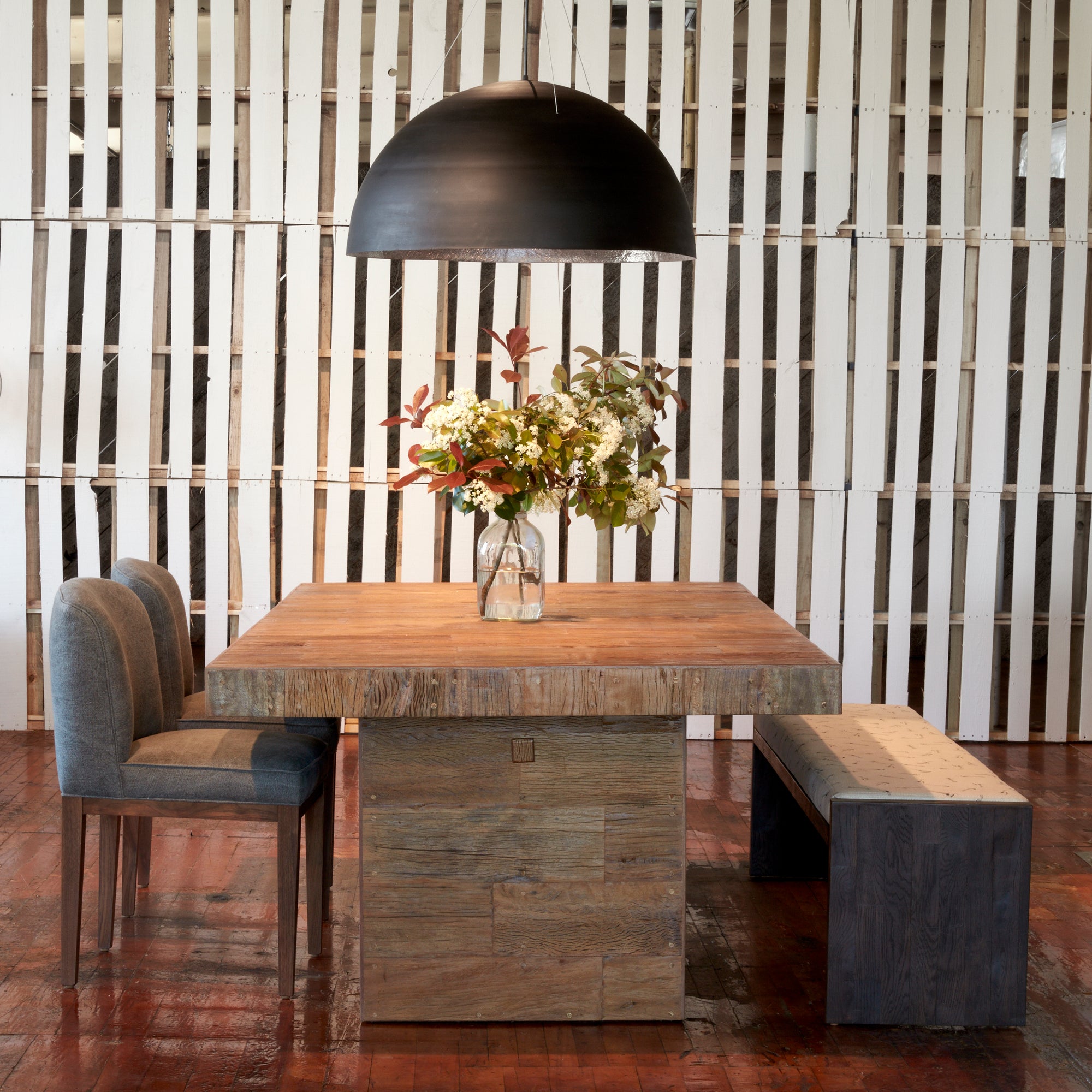  Reclaimed wood dining table shown with upholstered chairs in a grey fabric on one end and a bench for additional seating on the other end. Above dining table is the Bolle Pendant which is a circular pendant in a black finish.  