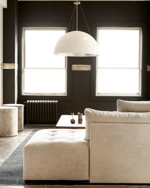  Tufted upholstered sectional in a neutral fabric placed in the center of a room that has black painted walls and two large windows. A white circular pendant hangs from the ceiling over a coffee table. Image is shot from the side profile.  