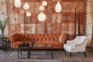  The Brook sofa is in a brown leather called Marvell Chesnut with a light colored chair on the right side. The room is colorful with a brown colored tapa cloth hanging in the background. There is a metal and glass coffee table and a cluster of white Jug lamps. Photographed in Marvell Chestnut. 