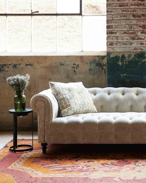  Daytime lighting of the Brook sofa in a Velluto Natural fabric, with a large window and a brick wall. There is an accent pillow and a Rotor side table to the left. The rug has a floral pattern in the red and orange tones. Photographed in Velluto Stone. 