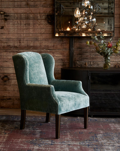 Chair in Velluto Aqua next to a dark credenza. In the background is a wood wall with a mirror and chandelier hanging. Photographed in Velluto Aqua.