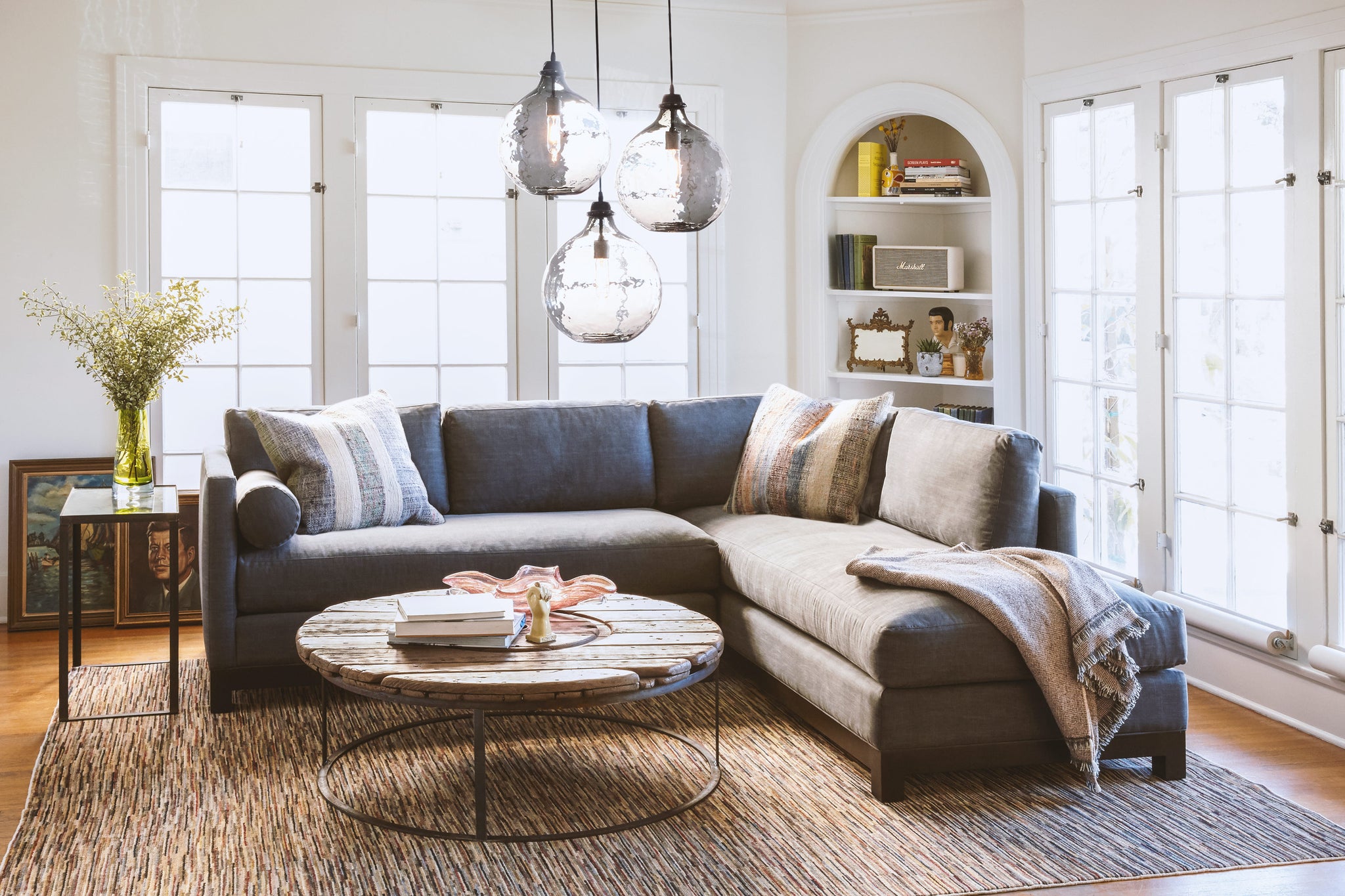  Cosmo Sectional sits in the center of a room next to a wood coffee table and glass side table. In the background are large windows with home decor items such as books and paintings. Above the sectional hangs 3 jug lamps. 