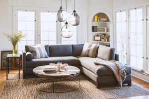  Cosmo Sectional sits in the center of a room next to a wood coffee table and glass side table. In the background are large windows with home decor items such as books and paintings. Above the sectional hangs 3 jug lamps. Photographed in Molino Slate. 