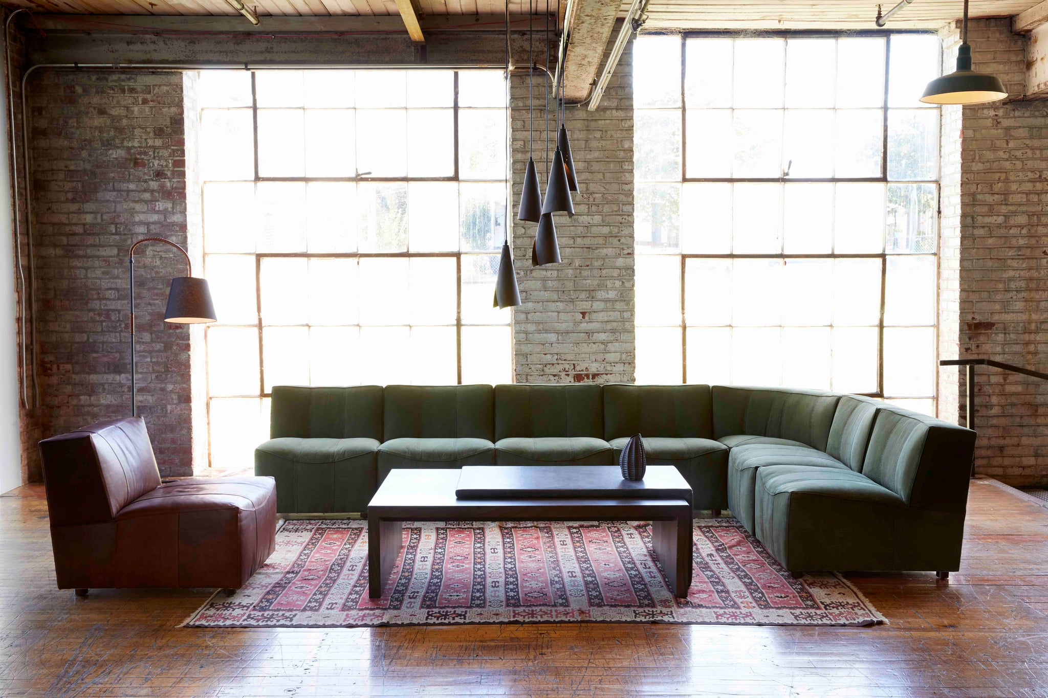  Century Sectional sits in the middle of a room with a wood coffee table and a leather chair. There is also a red patterned rug underneath the pieces. In the background is a brick wall with large windows. Above the pieces hangs multiple lighting lamps. Photographed in Hunter Green. 