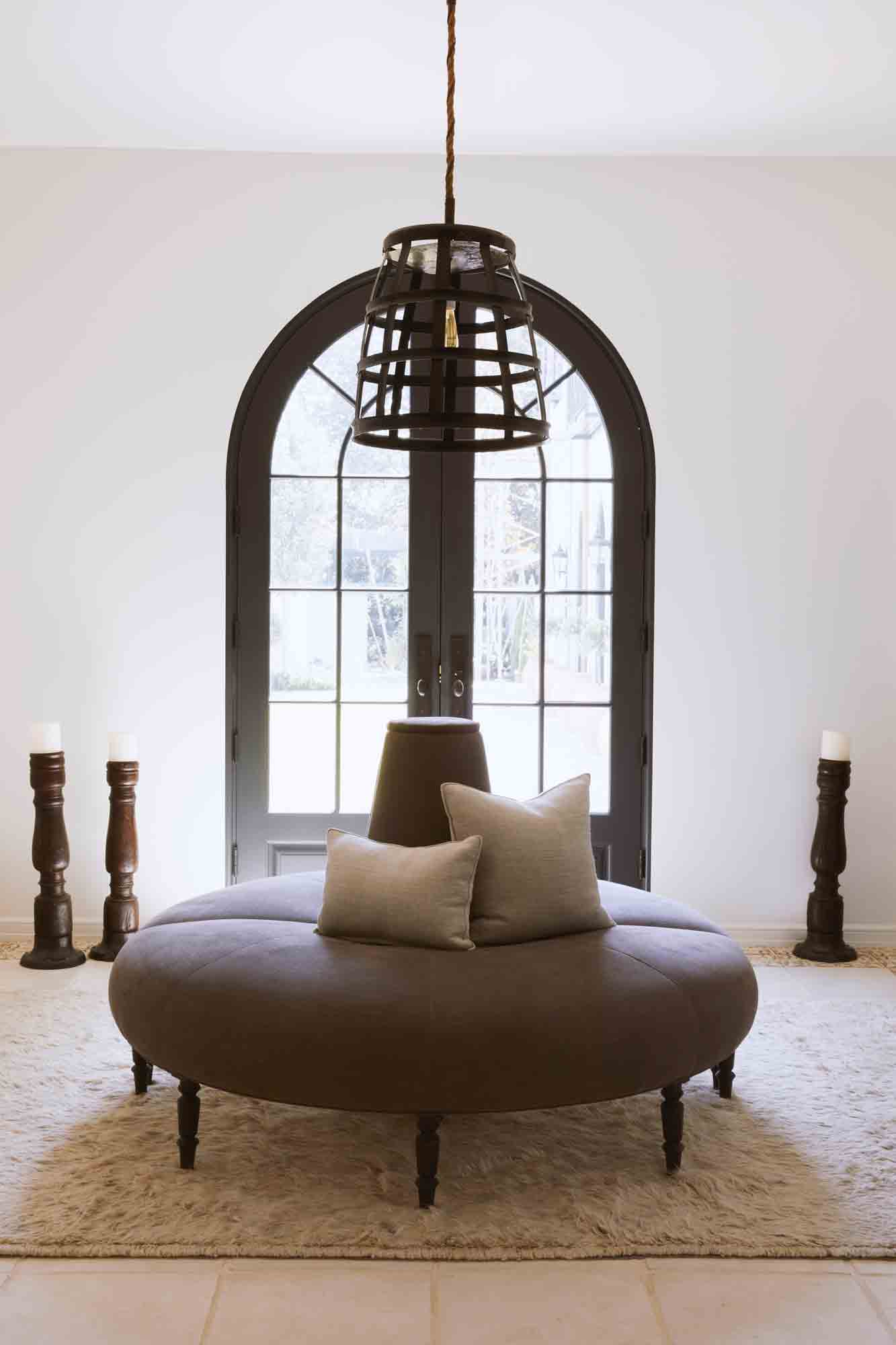  The Coin Sofa is in a dark grey linen, in a white foyer with a black metal chandelier and a double door with glass in the background. Photographed in Vintage Steel. 