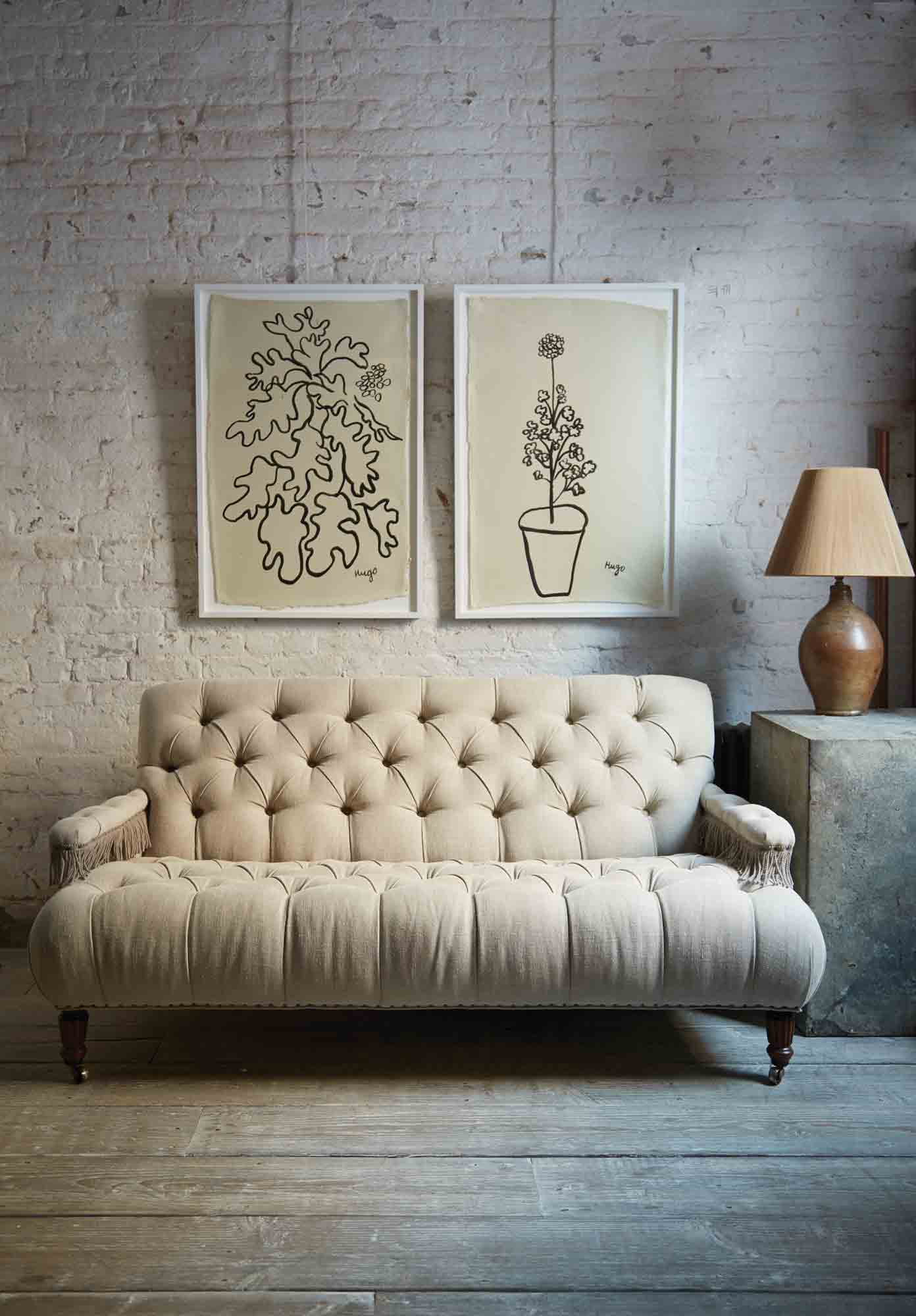  Tufted small scale sofa in a natural Vintage Flax linen with fringes on the arm rests. It is in a room wit daylight and a white brick wall and a wood floor. There are 2 pictures hanging on the wall and a table lamp on the right side. Photographed in Vintage Flax. 