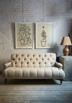  Tufted small scale sofa in a natural Vintage Flax linen with fringes on the arm rests. It is in a room wit daylight and a white brick wall and a wood floor. There are 2 pictures hanging on the wall and a table lamp on the right side. Photographed in Vintage Flax. 