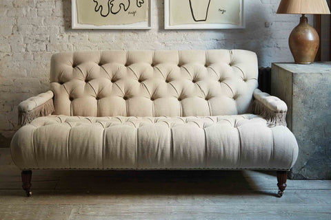Tufted small scale sofa in a natural Vintage Flax linen with fringes on the arm rests. It is in a room wit daylight and a white brick wall and a wood floor. Photographed in Vintage Flax.