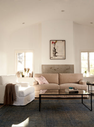  Large high ceiling living room with a sofa in beige linen and a white slipcovered chair on the left. A mirrored rectangular coffee table is in the middle. A painting is on the wall in the back of the room. Photographed in Brevard Burlap. 