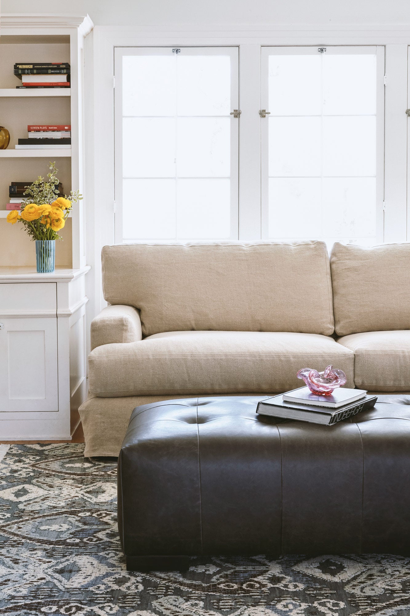  Daylight in a living room with a sofa in natural color linen in front of a window. A dark grey leather tufted ottoman sits in front of the sofa, on a blue, grey and white rug. There is a bookcase on the left side of the sofa. Photographed in Brevard Burlap. 