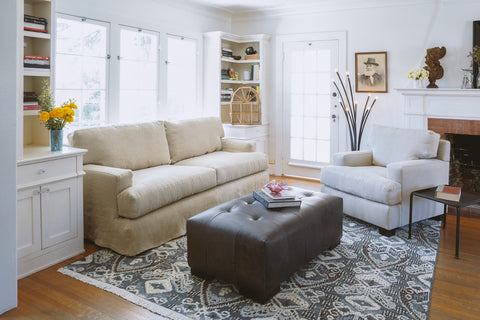 Daylight in a living room with a sofa in natural color linen in front of a window, an armchair is on the left side of the image in front of a fireplace. A dark grey leather tufted ottoman sits in the middle on a blue, grey and white rug. There are 2 bookcases on each side of the sofa. Photographed in Brevard Burlap.