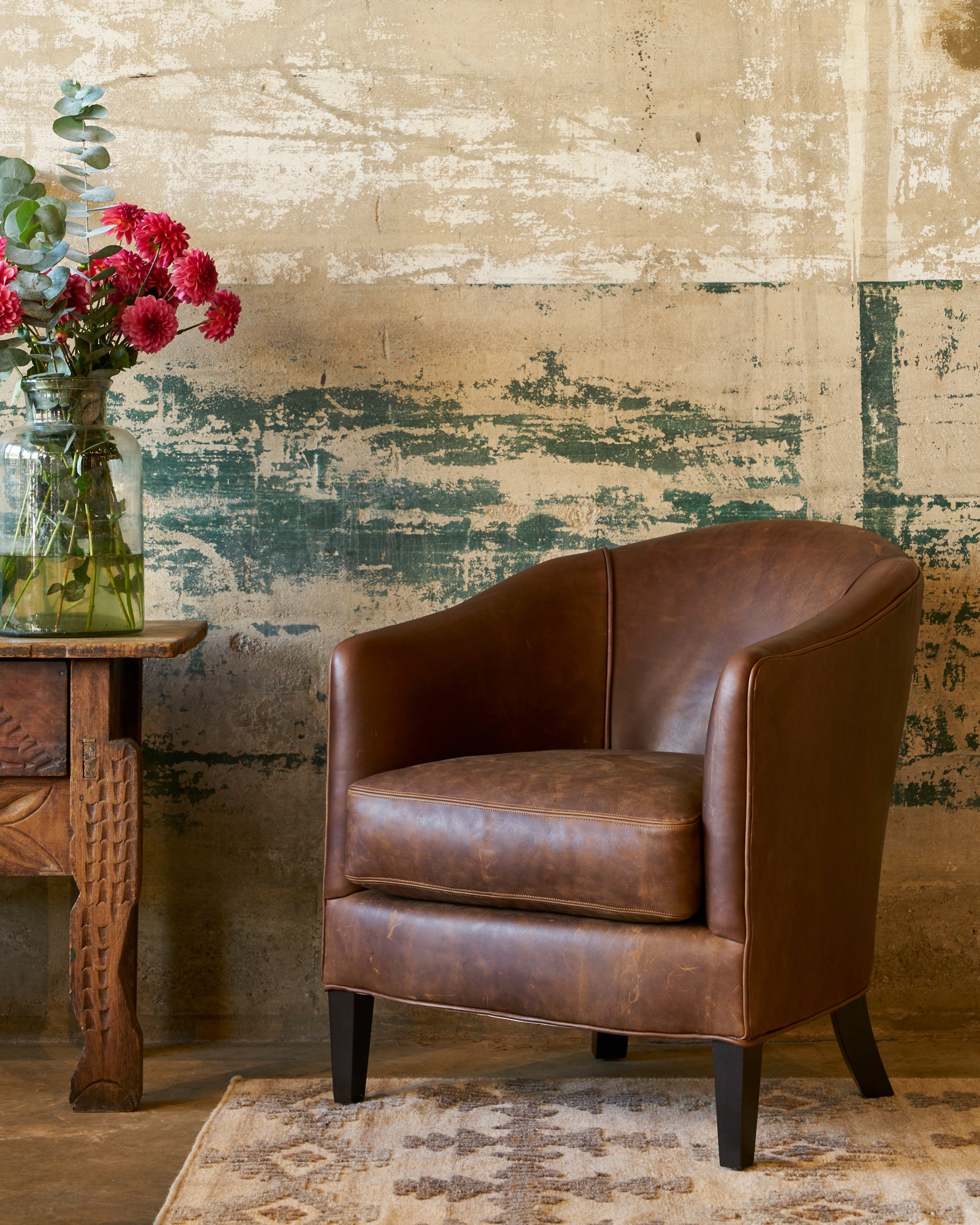  Spur Chocolate Crescent Chair in brown leather without nailheads in front of a patinaed cement wall with a vintage wood table on the left and red flowers on top. Photographed in Spur Chocolate. 