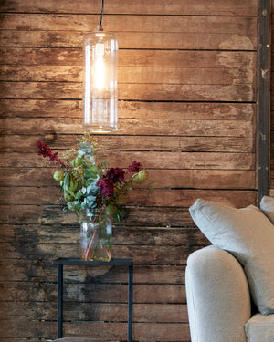  Cylinder lamp in clear hanging against a wood wall above a flower arrangement. On the right, a detail of a sofa.  