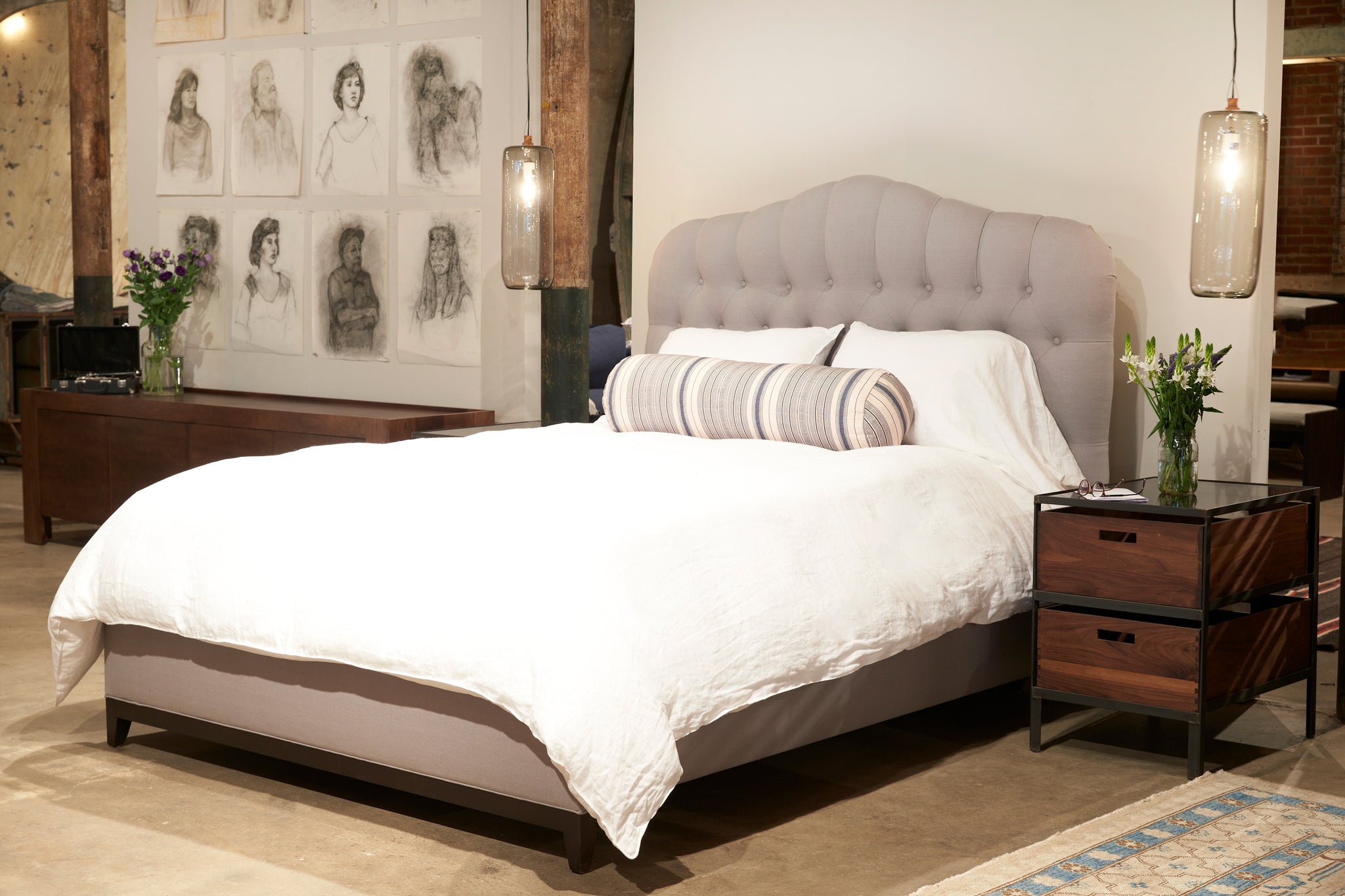  Tufted upholstered bed in a light grey fabric with white bedding and an accent pillow in striped fabric. Bed has a wood nightstand with glass top and there are two cylinder pendants hanging on either side of the bed in smoke finish.  