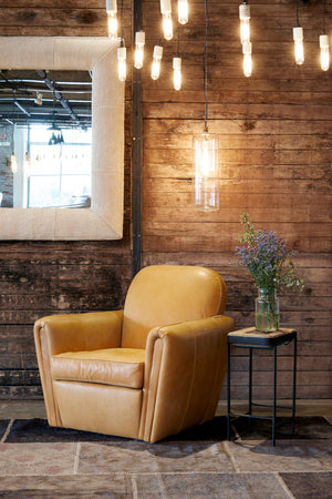  Upholstered club chair in a mustard leather there is a small side table along side the chair and a cylinder pendant hanging above side table in clear finish. Chair is placed against wood wall and there is an upholstered wall mirror hanging on the wall.  