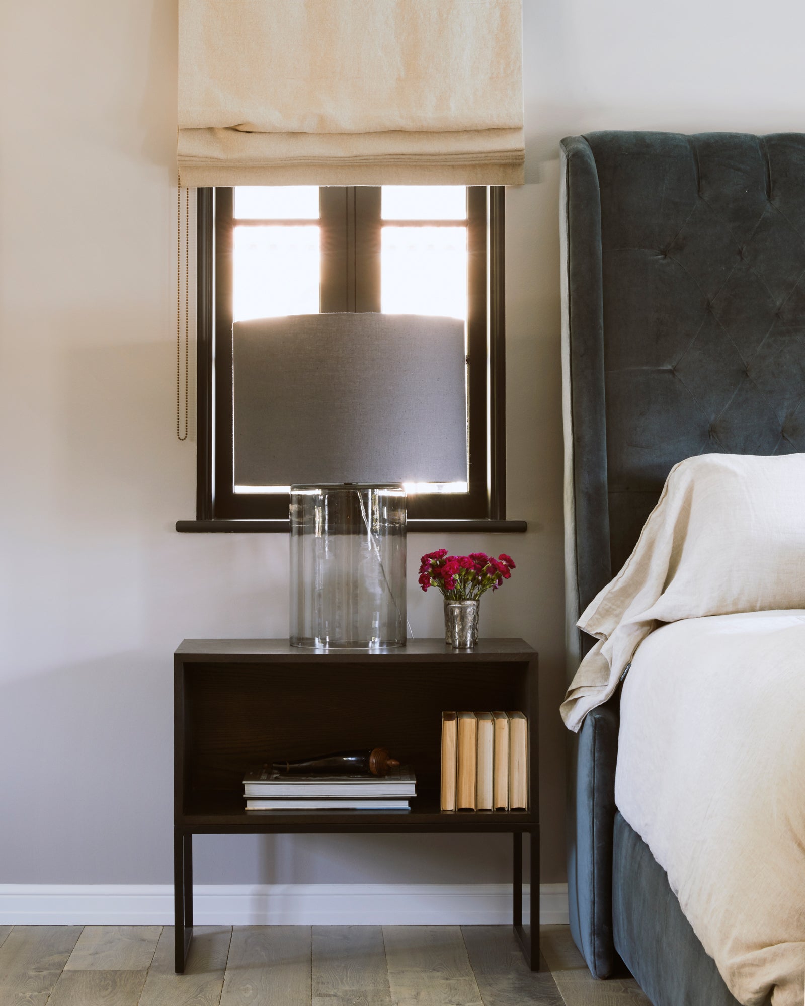  Detail shot of bedroom showing the corner of a upholstered bed in dark grey fabric and white bedding. There is a wood nightstand alongside the bed and a cylinder table lamp on top in a smoked finish and has a grey fabric lampshade.  