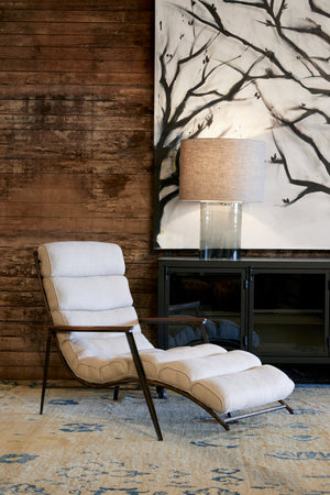  Curved upholstered lounge chair against a wood wall with a metal credenza and black and white art work on the wall. There is a cylinder table lamp in smoked finish and has a fabric lamp shade in a neutral color. 