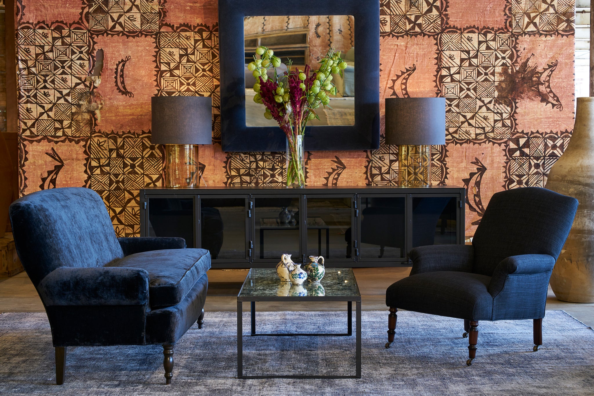  Isaac Credenza in flat black finish in front of a wall with colorful motif. Wall mirror upholstered in blue velvet and black velvet sofa in front with a dark blue chair. 