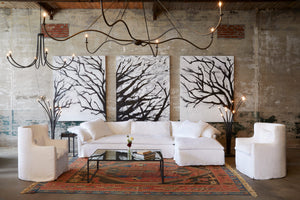  Donato 2pc Sectional in Otis White next to two chairs and a glass coffee table. Underneath the sectional is a patterned rug. They are also floor lamps next to the sectional. In the background are large paintings of tree branches. Above the sectional are multiple chandeliers. Photographed in Otis White. 