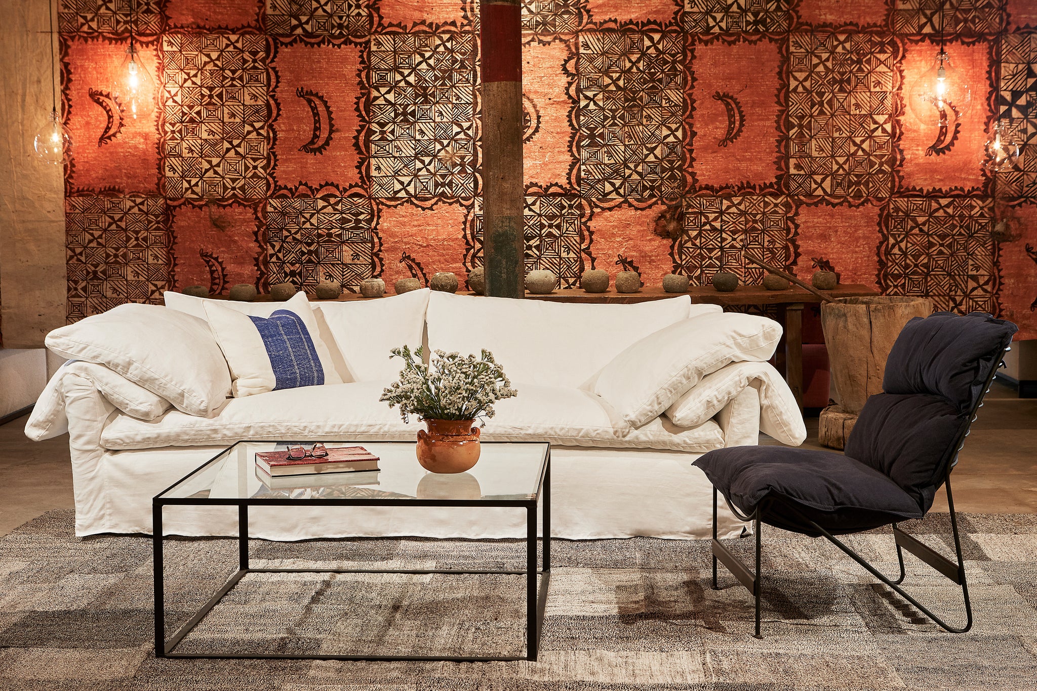  The white linen slipcovered sofa is in front of a vintage Tapa Cloth with orange and brown graphics. The chair on the right side has a metal frame and a dark blue cushion. The coffee table is in metal with a glass top. Photographed in Logan White. 