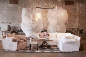  Donato 2 arm Sectional in Otis White next to a leather ottoman and a metal coffee table. In the background are two art pieces that resemble faces. A chair is next to the sectional. Underneath the sectional is a grey, diamond patterned rug. Photographed in Otis White. 