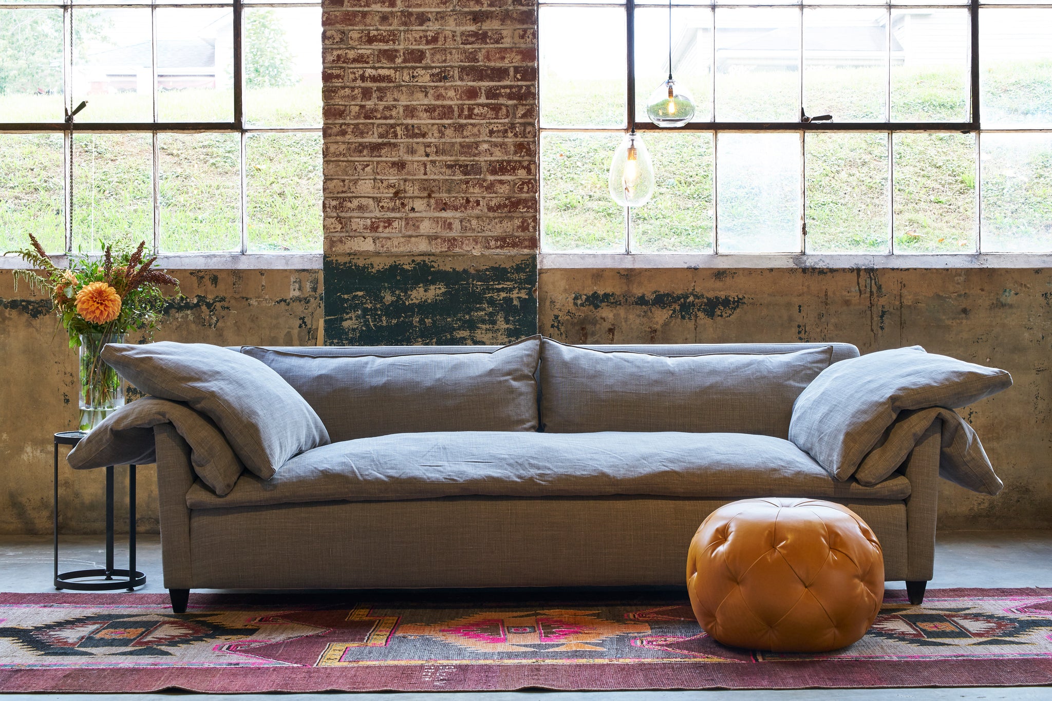  Rye Warm Grey Donato Sofa upholstered in a brown linen in front of large windows and an orange leather pouf in front. Photographed in Rye Warm Grey. 