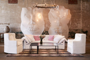  The white slipcovered sofa is in a large room with 2 gigantic white face mask sculptures on the wall. There are 2 white slipcovered wing chairs on each side and a metal and glass coffee table in the middle. Photographed in Otis White. 