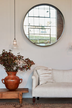  Daylight in a room with the sofa against a white wall with a round mirror and a Morse Pendant light. There is a wood table on the left with a terracotta pot on it and branches inside. Photographed in Vintage Hemp. 
