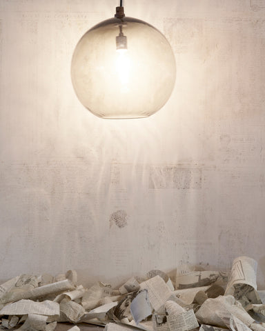Detailed shot of Globe Pendant in smoke hanging against a white wall and torn newspapers on the floor. 