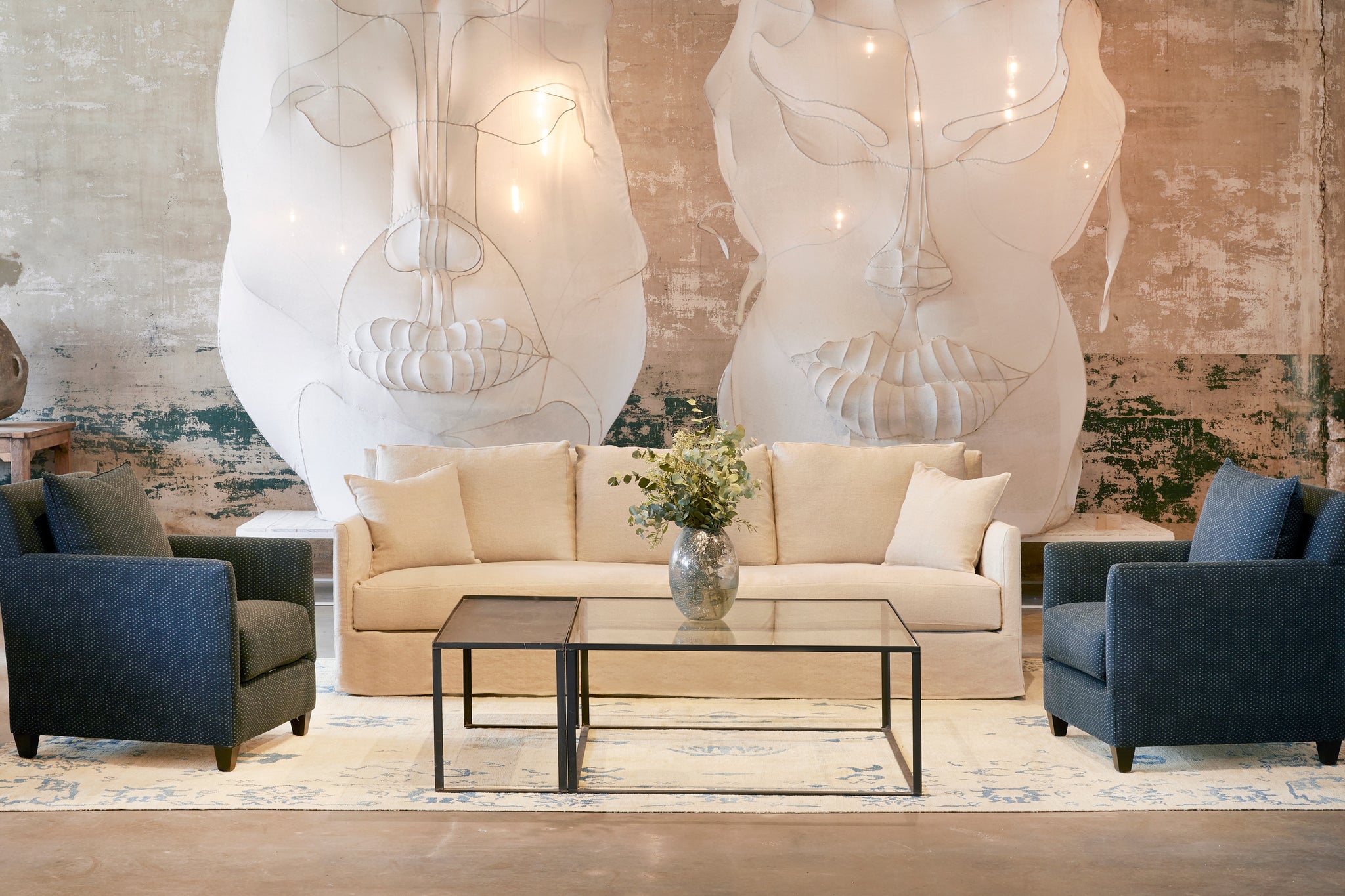  The 96" slipcovered sofa is  in Brevard Burlap, in a large room with 2 white face mask sculptures on the wall. There are 2 upholstered chairs on the side, in dark blue fabric with small dots and a metal and glass coffee table in front. Photographed in Brevard Burlap. 